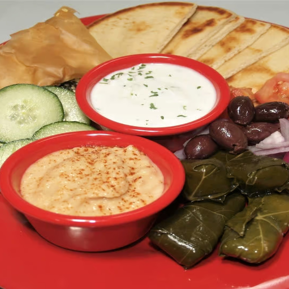 A Greek appetizer platter with hummus, tzatziki, pita bread, dolmades, cucumbers, olives, and tomatoes.