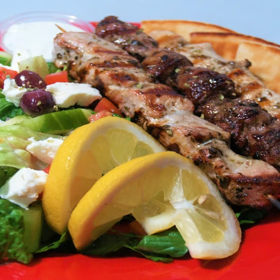 Grilled Greek souvlaki skewers with lemon wedges, salad, and pita bread on a red plate.